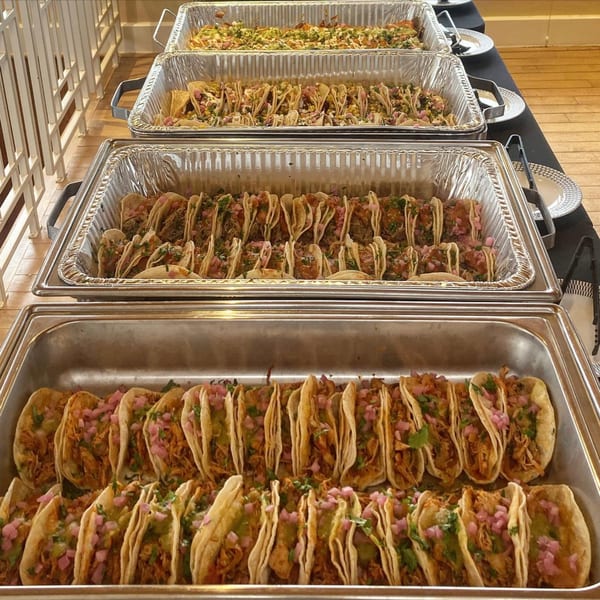 Four large catering trays filled with various La Prensa tacos, including chicken and pork.