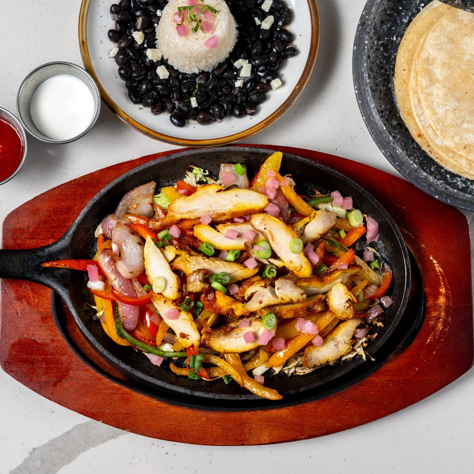 Chicken fajitas sizzling in a cast iron skillet with rice, black beans, tortillas, and sauces.